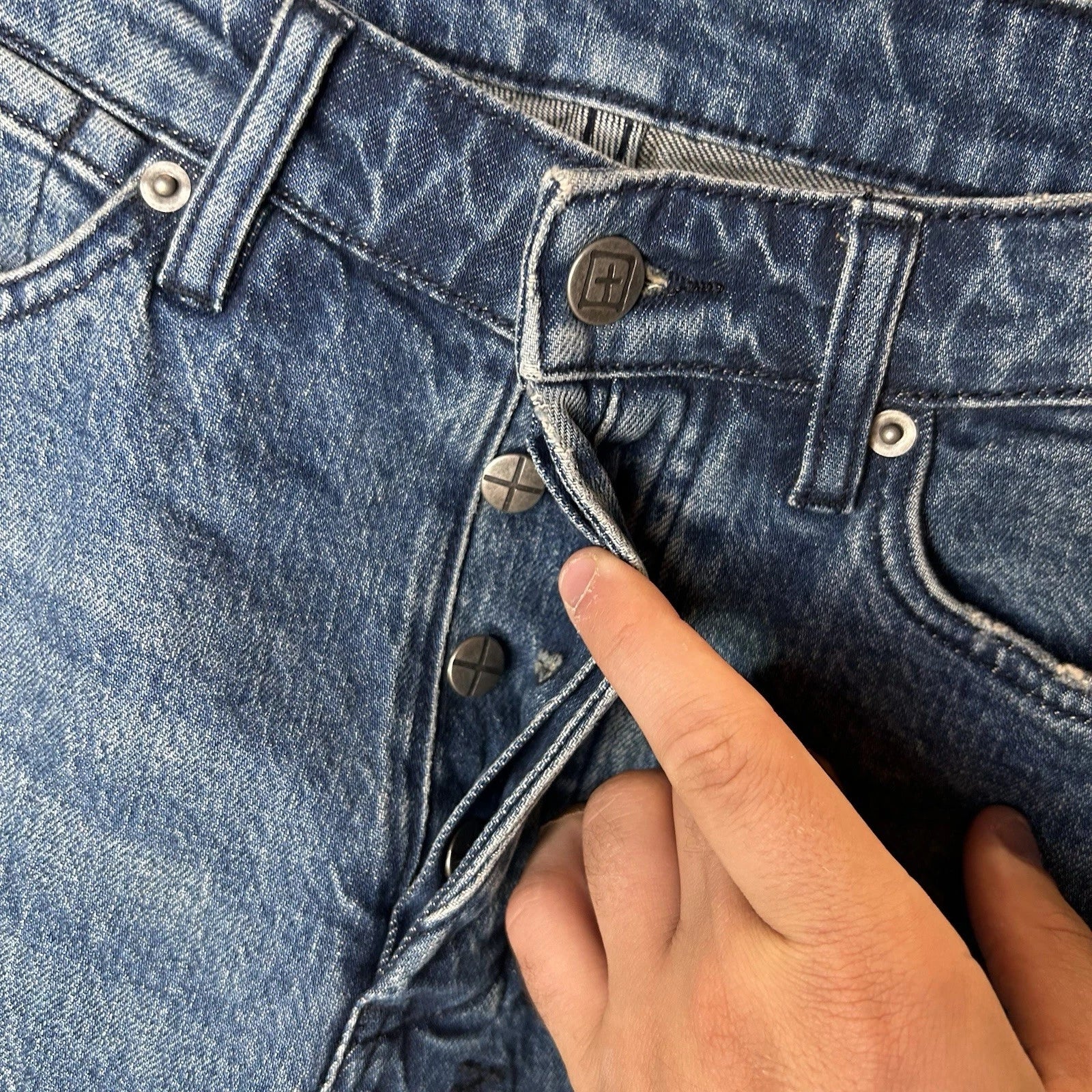 KSUBI blue denim jeans button fly, hand revealing signature cross buttons.